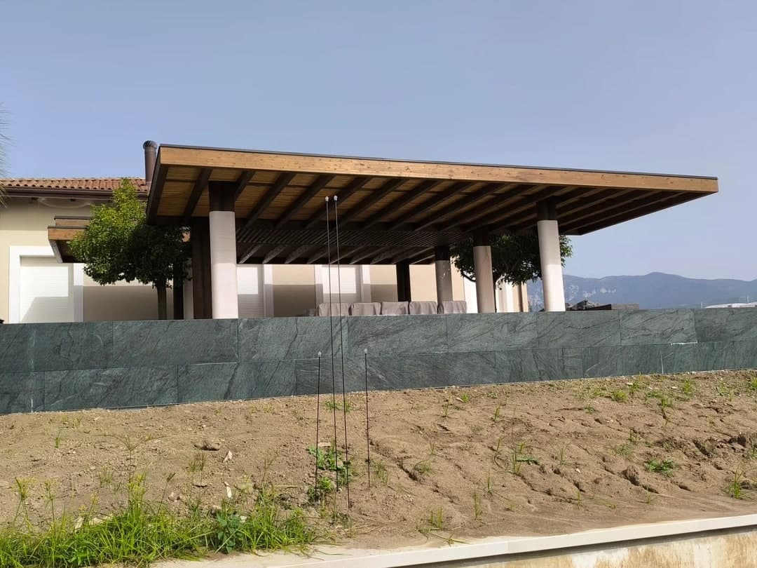 La pergola vista dal basso, oltre il muro della terrazza rivestito di lastre scure: il tetto sottile e largo sulle colonne bianche; dietro, i monti.