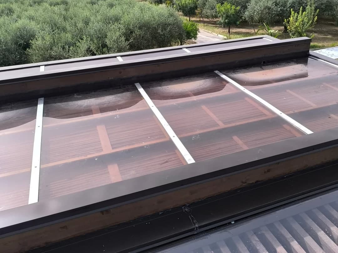Il tetto della pergola visto dall’alto: lastre ondulate trasparenti color bronzo, i coprigiunti bianchi fra una lastra e l’altra, il bordo testa di moro; attorno un uliveto.