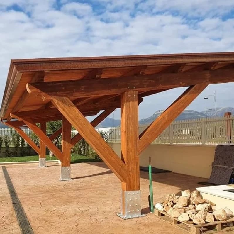Da vicino, un pilastro in legno lamellare su una scarpa d’acciaio, con le due saette che si aprono a Y sotto la trave; accanto un bancale di pietre, sotto un cortile in cemento stampato.