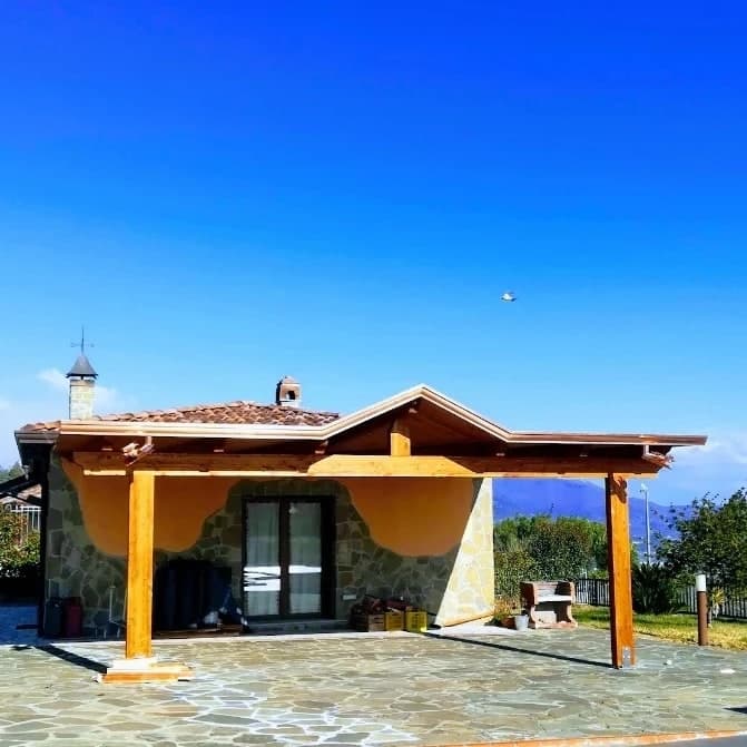 Una casa a un piano in pietra, con l’intonaco ocra in alto e il tetto in coppi; davanti, un portico in legno chiaro su due pilastri, con un frontone a capanna sopra la portafinestra.