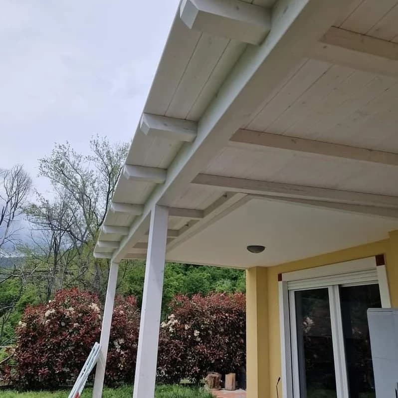 La tettoia da sotto, verso il giardino: pilastri, trave, travetti e tavolato verniciati di bianco; dietro, le siepi rosse e la collina.