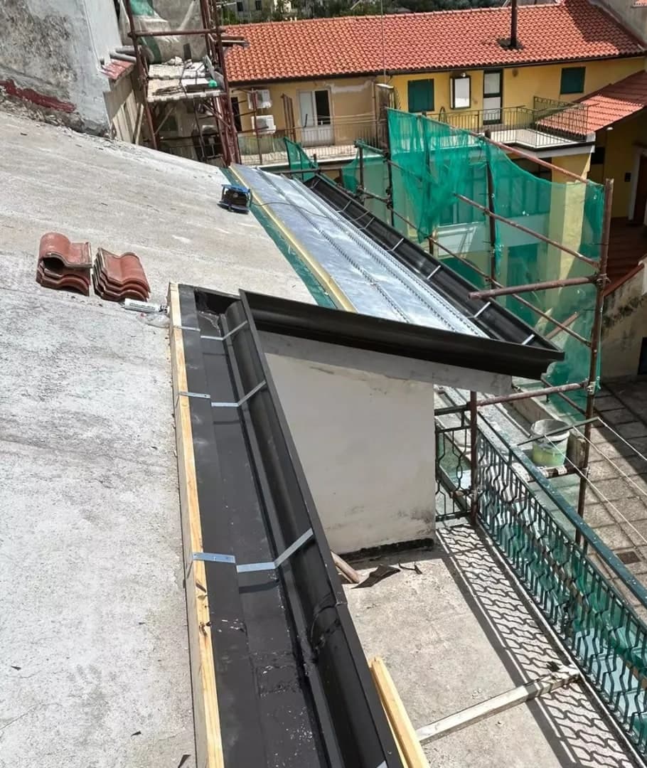 Dall’alto, il canale di gronda testa di moro sul bordo del tetto piano, con le staffe zincate di traverso e la tavola di legno; più in là il muro bianco del corpo che sporge, la terrazza con la ringhiera verde, il cortile e le case in coppi.