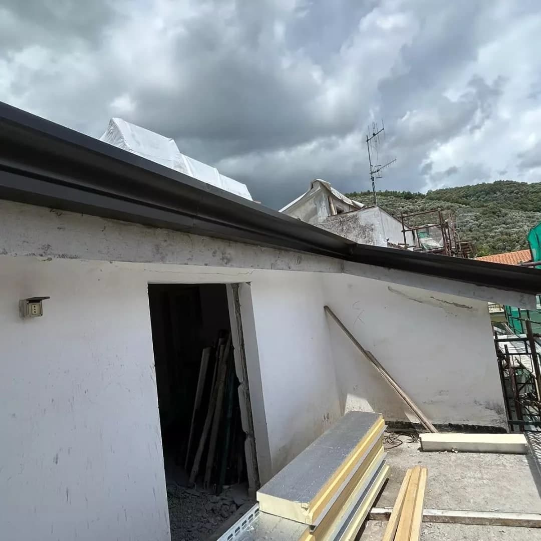 Dalla terrazza, il corpo dal muro bianco con la porta e, in cima, la faccia scura del canale; dietro, le colline; per terra i pannelli coibentati accatastati.