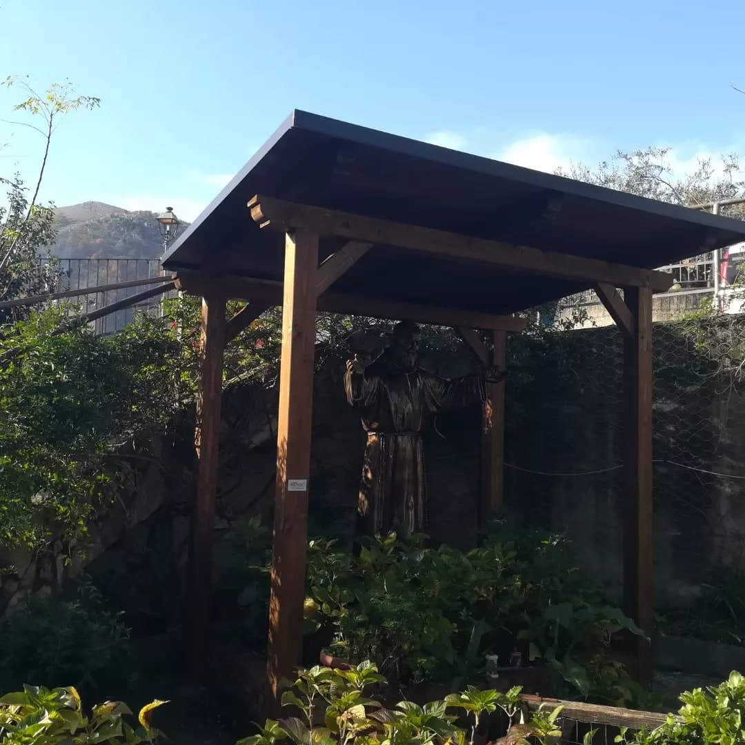 La piccola tettoia in legno nel giardino, dal davanti: quattro pilastri, le travi con le teste sagomate, le saette; sotto, una statua fra le piante; dietro, il muro e i monti.