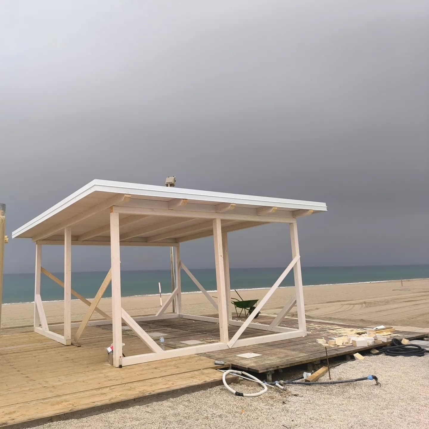 La struttura in legno bianco sulla pedana, con il mare dietro: tre pilastri per lato, le travi in basso e in alto, le croci di controvento, il tetto piano con il bordo bianco.
