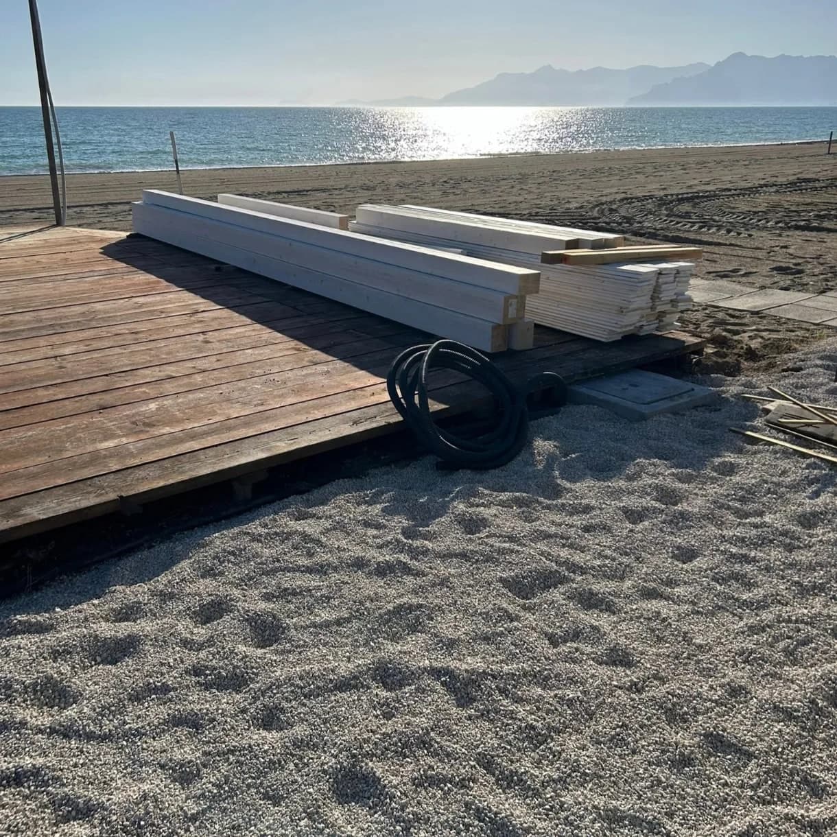 Prima del montaggio: le travi verniciate di bianco posate sulla pedana, la sabbia, il mare e la costa.
