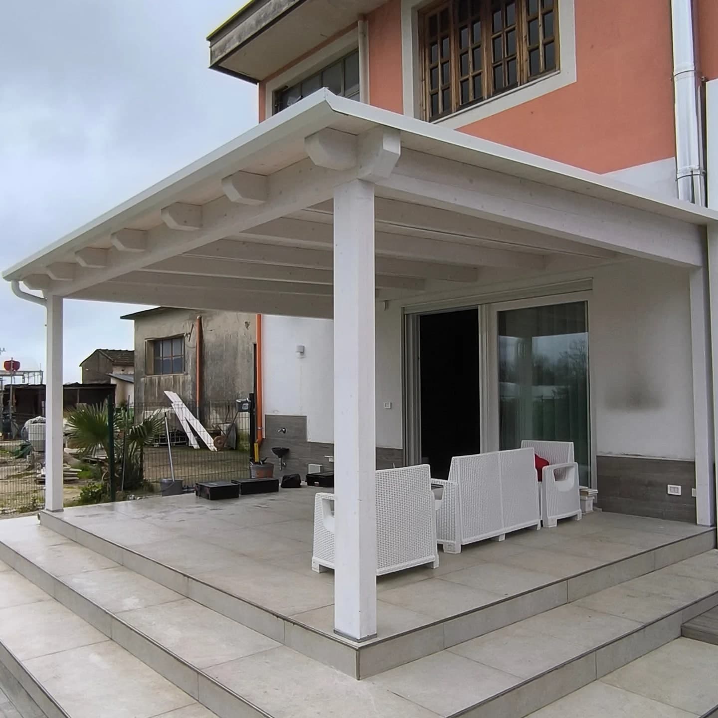 La tettoia bianca di tre quarti dall’angolo davanti a destra: la gronda lungo il davanti, le teste sagomate dei travetti, i pilastri sul patio rialzato; sopra, il primo piano rosa della casa; a sinistra, un rustico e il giardino.