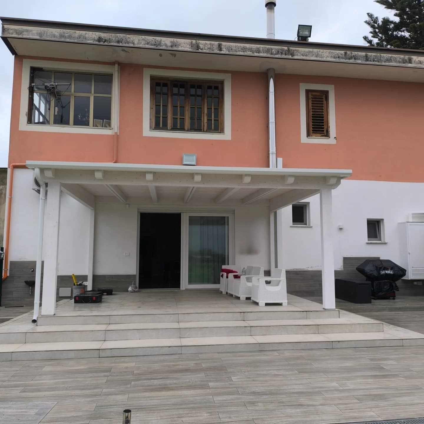Il retro della casa da lontano: il piano terra bianco con lo zoccolo scuro, il primo piano rosa con le finestre dai telai di legno, il tetto piano con lo sporto grande; sotto, la tettoia bianca sul patio con i gradini.