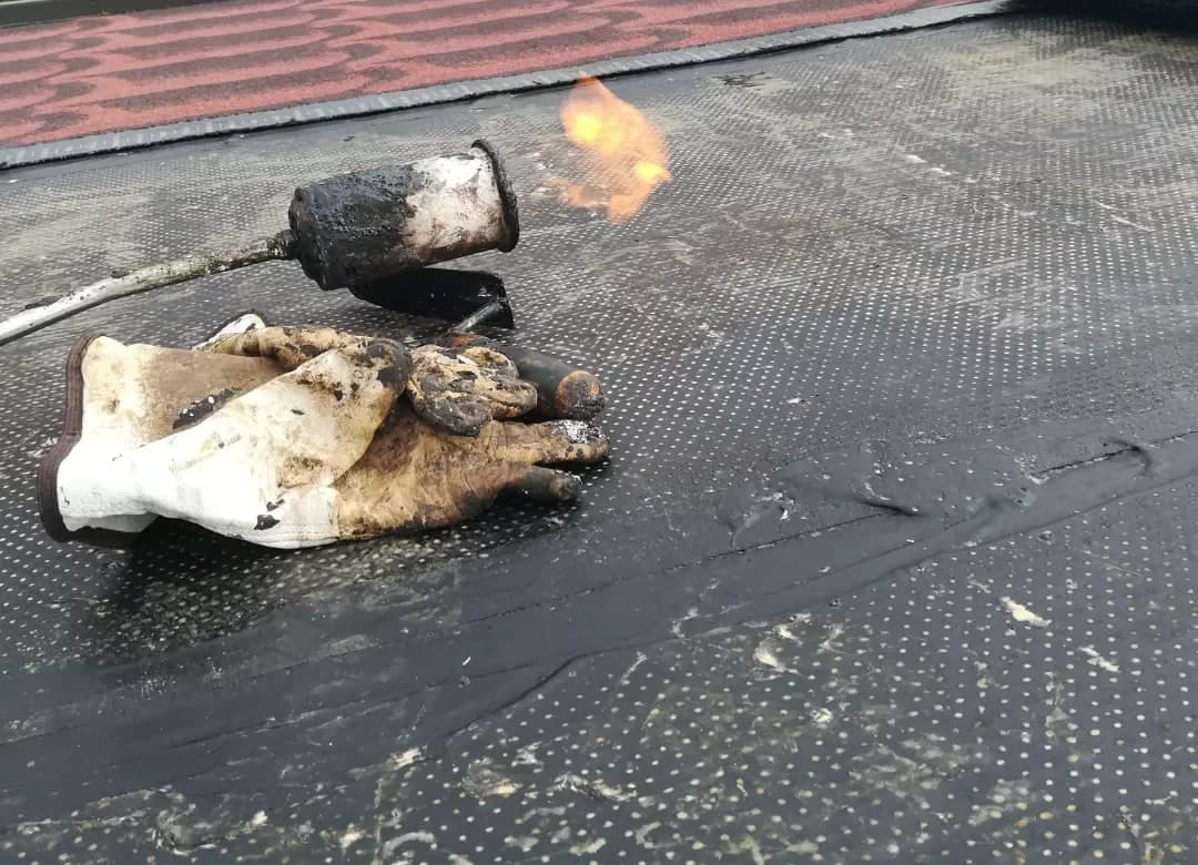 La posa: sul primo strato nero la guaina stampata, posata a fiamma; il cannello e i guanti da lavoro sopra il primo strato, il rotolo accanto.