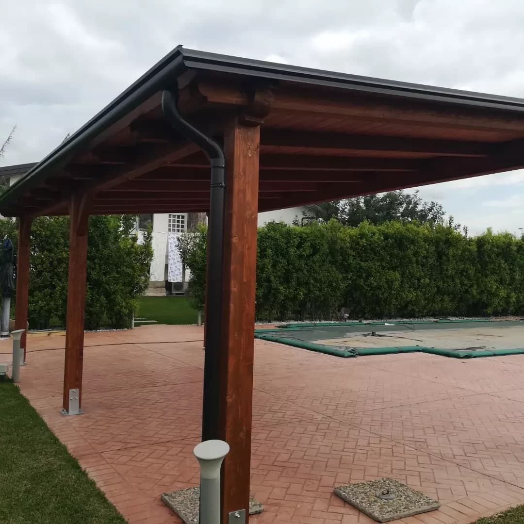 La tettoia in legno tinto mogano sul pavimento a spina di pesce, accanto alla piscina coperta dal telo: lungo il lato basso la gronda scura, e dal suo capo il pluviale scende con il collo d’oca lungo il pilastro d’angolo; sotto, i travetti e il tavolato.