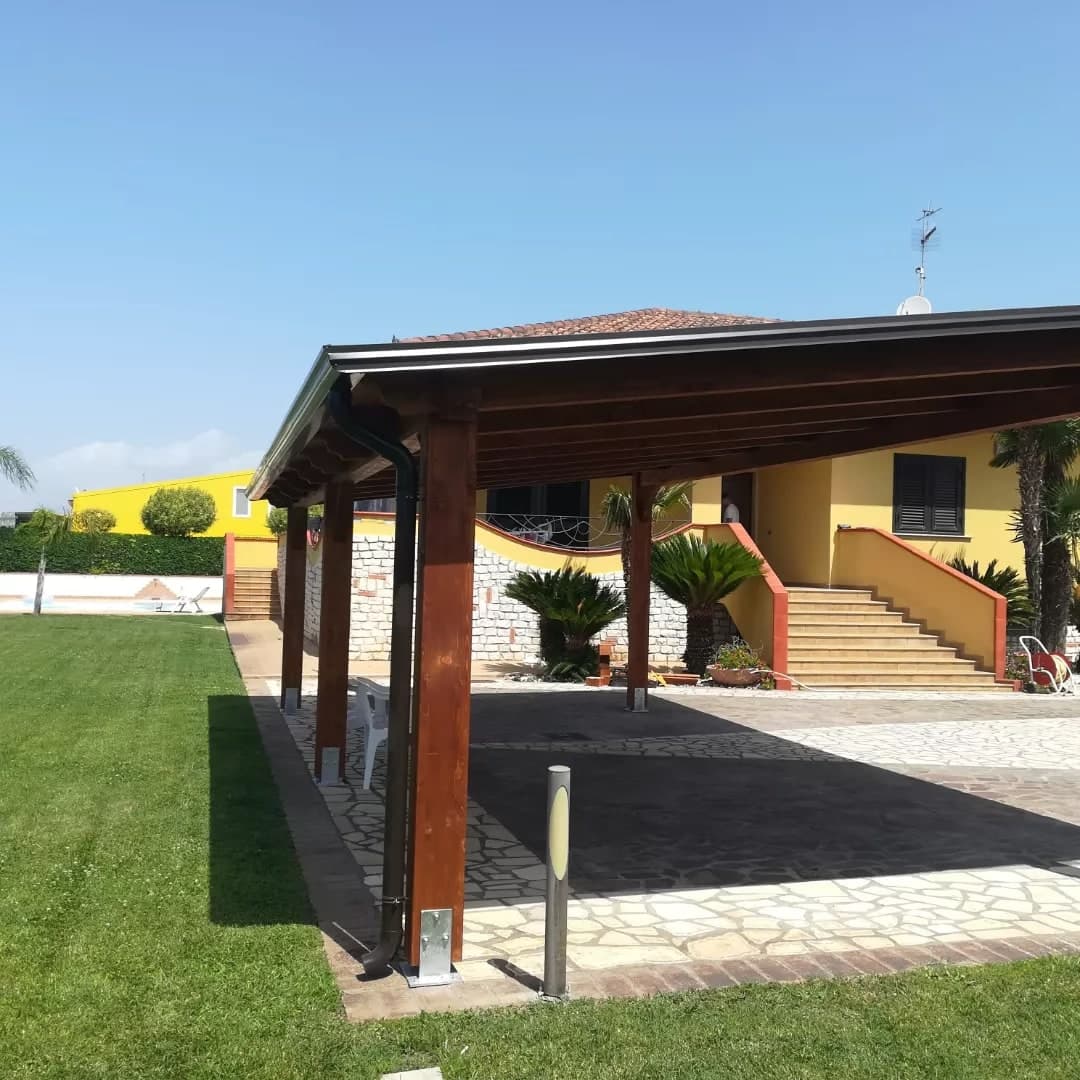 L’angolo della tettoia: il pilastro su una scarpa d’acciaio, la gronda testa di moro lungo il bordo e il pluviale che scende lungo il pilastro; accanto un paletto luminoso.