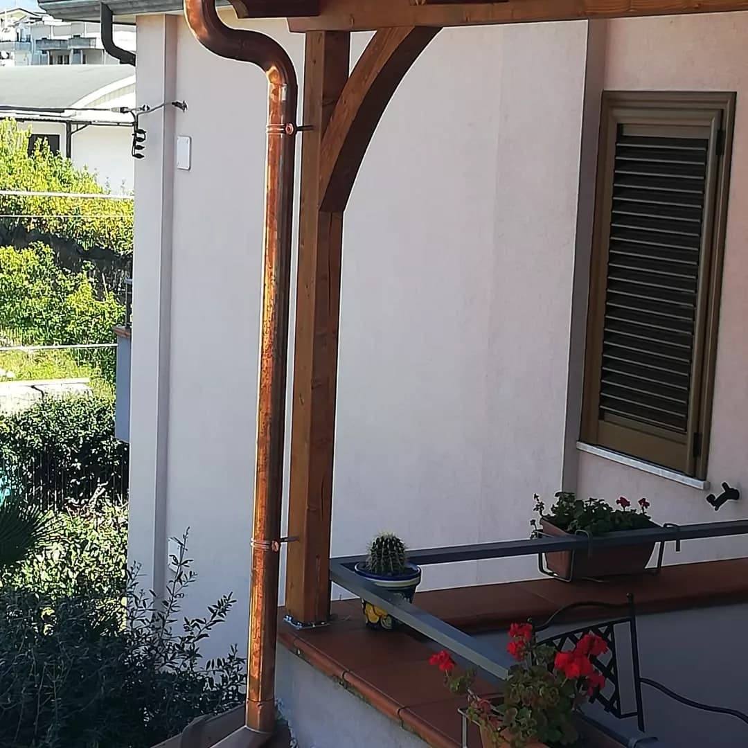 Il pluviale in rame nuovo che dal collo d’oca scende lungo il pilastro in legno della tettoia, tenuto da due collari, accanto alla mensola curva; dietro, il muro con una finestra dalle persiane chiuse e il balcone con i vasi.