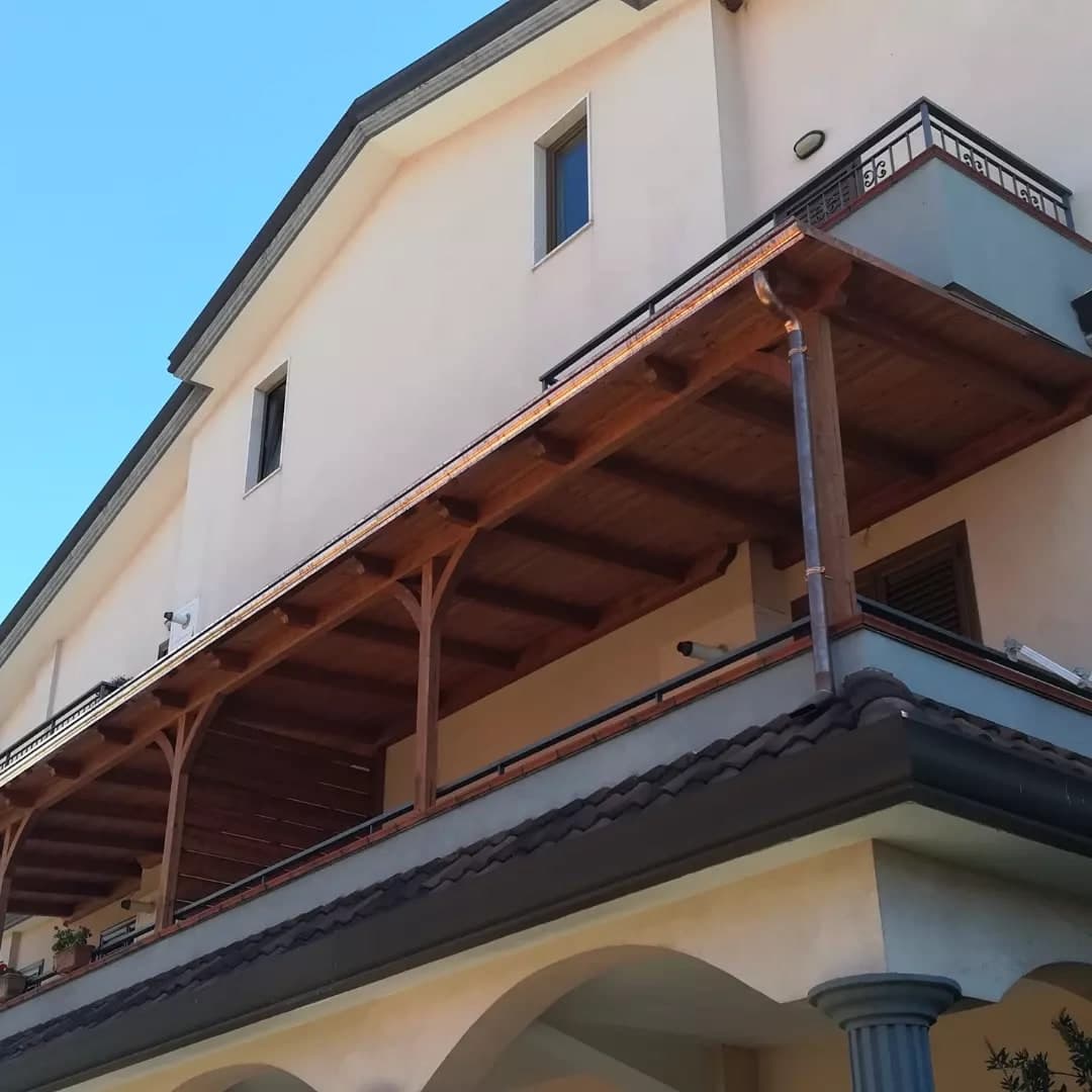 Da sotto, la villa crema con la tettoia in legno sul balcone del primo piano: i pilastri con le mensole curve, i travetti con le teste sagomate, la gronda in rame lungo il bordo e, in fondo, il pluviale in rame con il collo d’oca giù lungo il pilastro; sotto, la falda in coppi scuri sopra il portico ad archi.