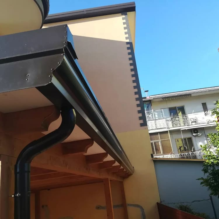 Gronda e pluviale scuri sotto un tetto in legno: la curva del pluviale scende lungo il pilastro.
